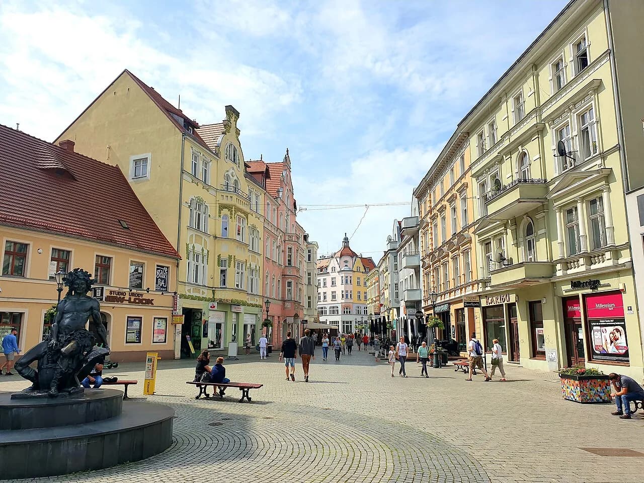A wide-angle view of a large, open town square with historical buildings on both sides and a fountain statue of a male figure grappling with a sea creature in the foreground.