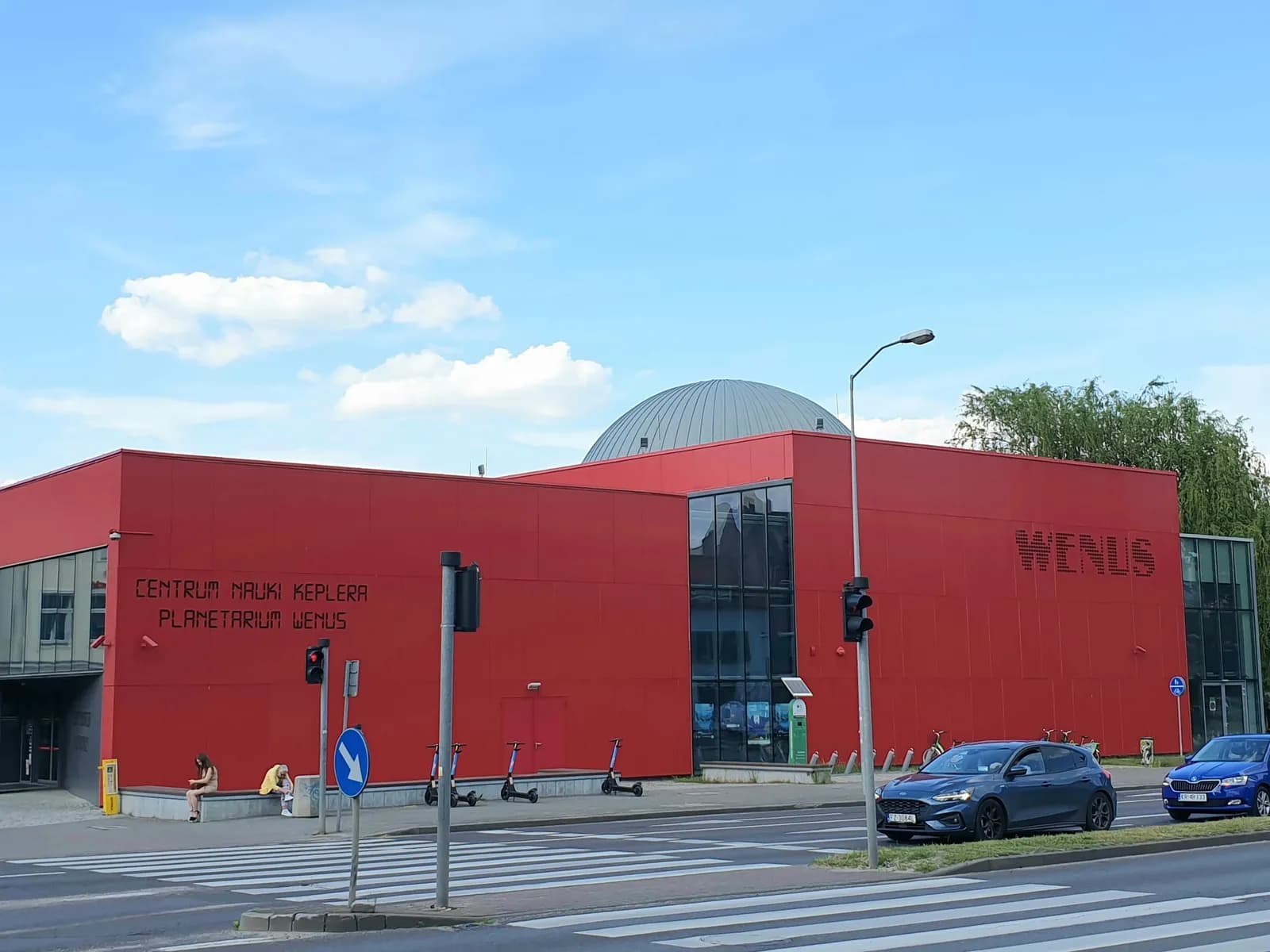 A modern building with a vibrant red facade and dark windows, featuring the text "CENTRUM NAUKI KEPLERA PLANETARIUM WENUS" on its wall under a blue sky.